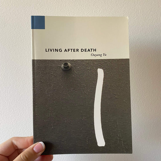 A hand is holding a paperback book titled 'Living After Death' by Ouyang Yu. The book has a white and blue cover with a minimalist design.