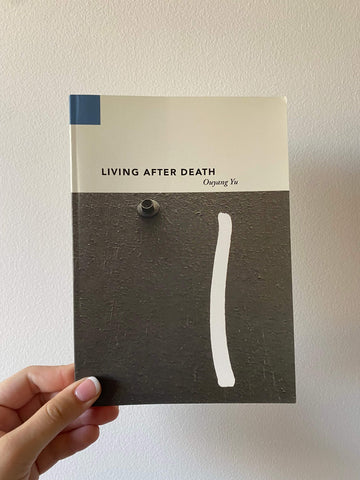 A hand is holding a paperback book titled 'Living After Death' by Ouyang Yu. The book has a white and blue cover with a minimalist design.