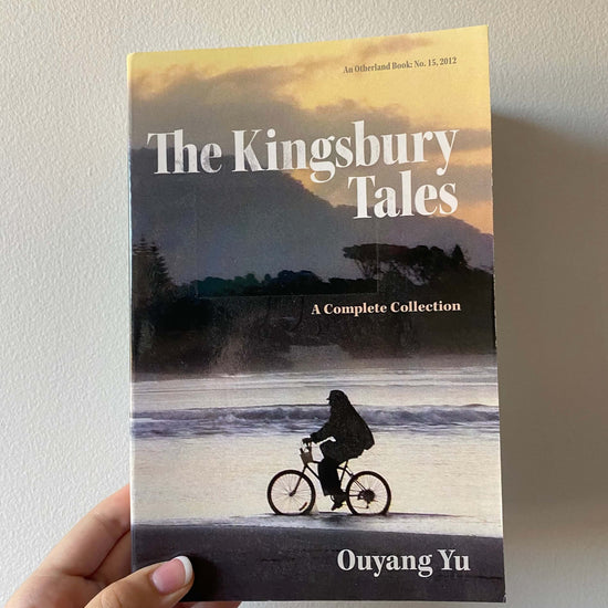 A hand holding a book titled 'The Kingsbury Tales: A Complete Collection' by Ouyang Yu, with a silhouette of a person on a bicycle on the cover.