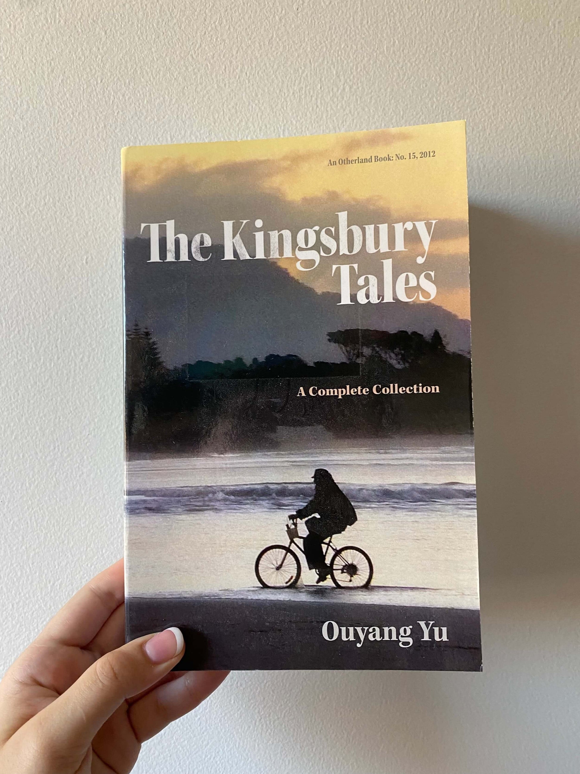 A hand holding a book titled 'The Kingsbury Tales: A Complete Collection' by Ouyang Yu, with a silhouette of a person on a bicycle on the cover.