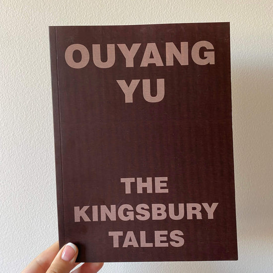 A hand holding a book titled 'The Kingsbury Tales' by Ouyang Yu with a plain brown cover.