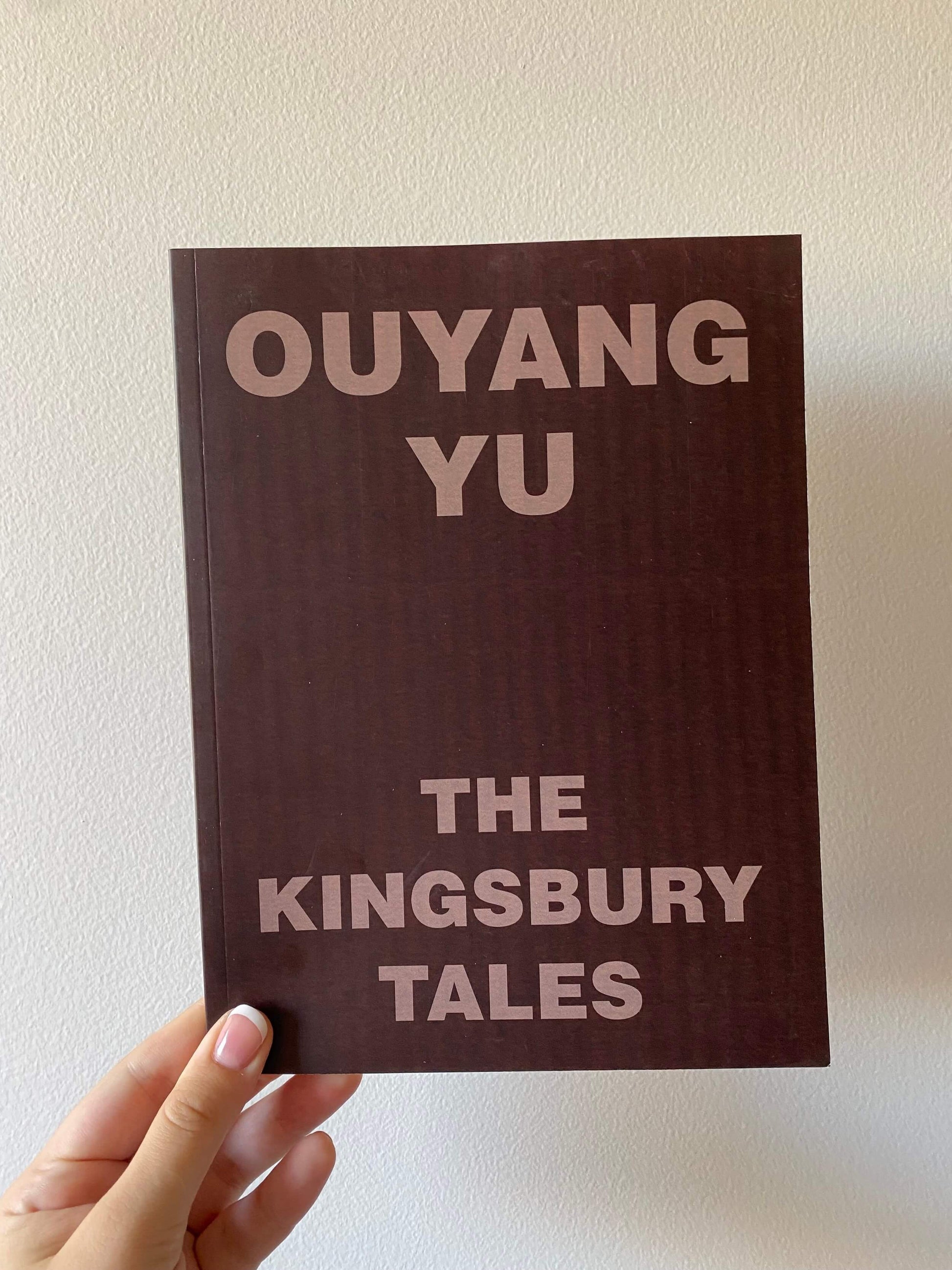 A hand holding a book titled 'The Kingsbury Tales' by Ouyang Yu with a plain brown cover.
